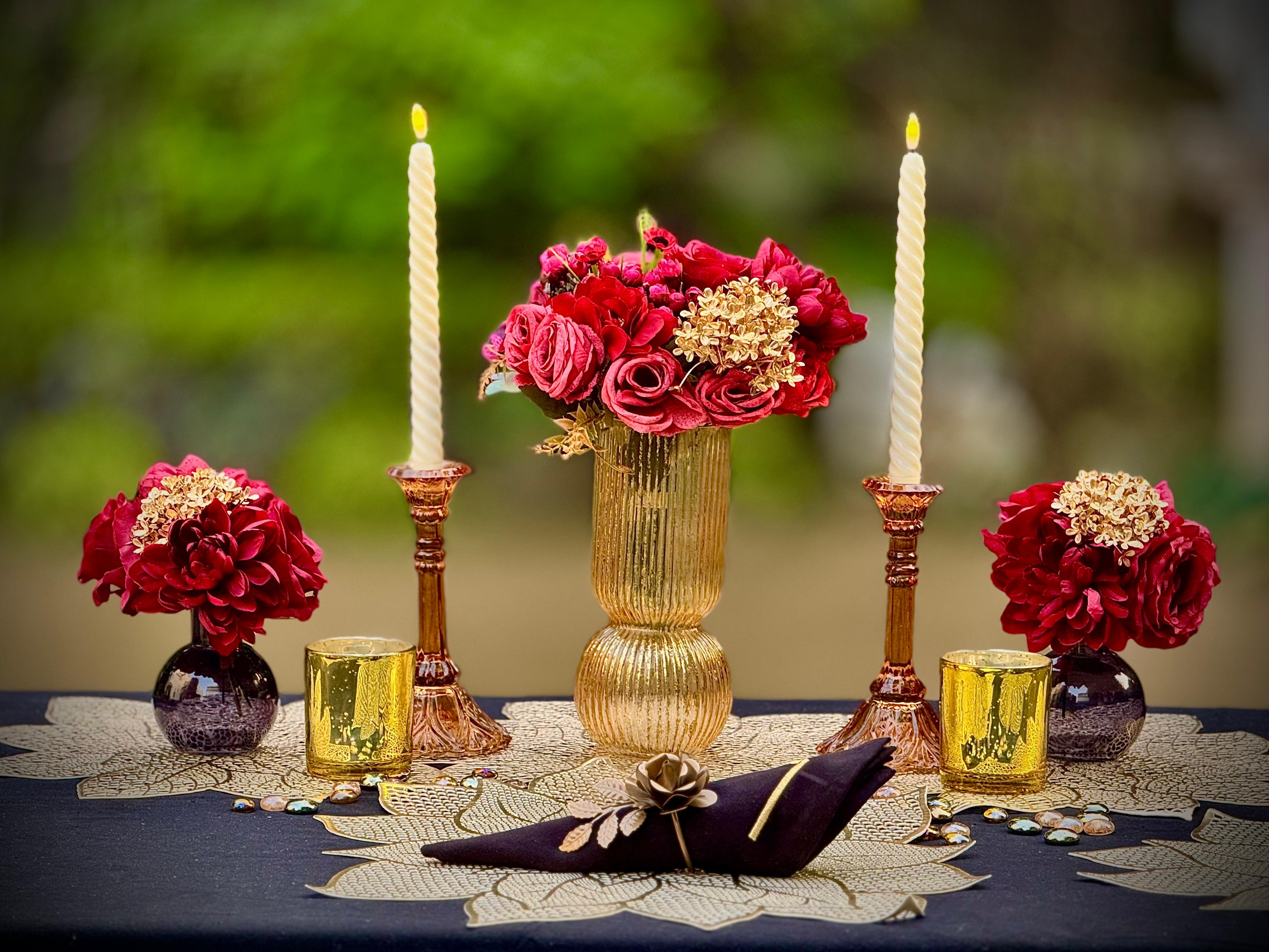 A table setup featuring a gold center vase with red and golden florals, surrounded by amber candle stands with taper candles, and embroidered napkins with flower napkin rings. The table has a dark cloth and is adorned with golden flower-shaped mats.